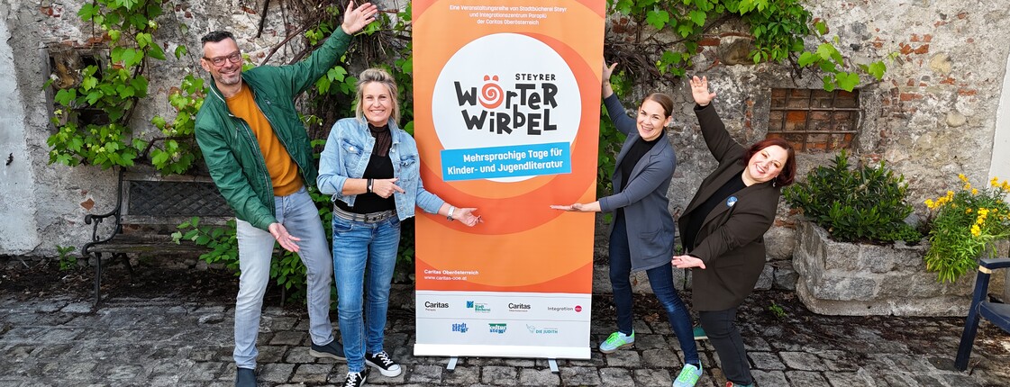 Vier Personen zeigen auf ein oranges Plakat mit der Aufschrift: Steyrer Wörterwirbel